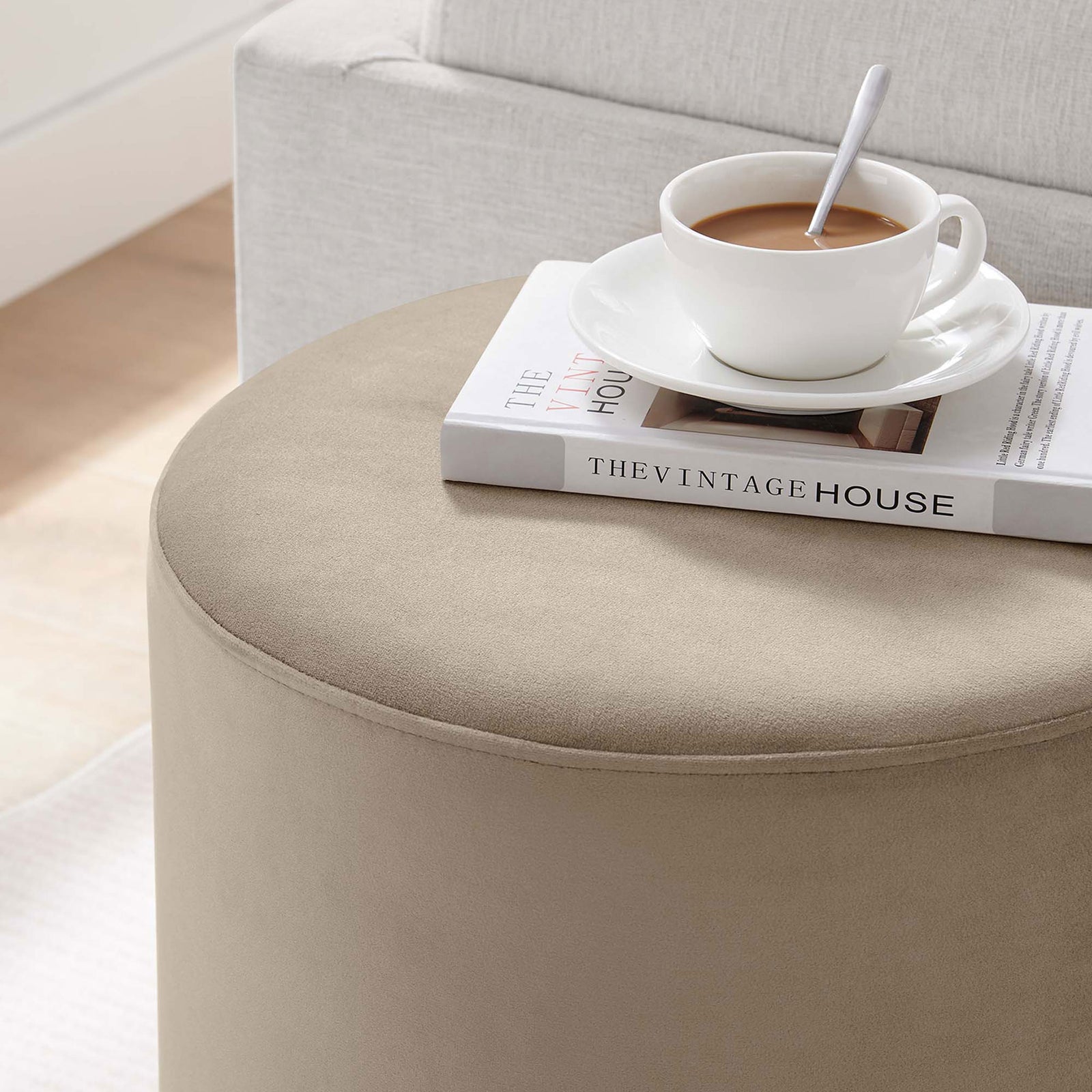 Round taupe ottoman used as a chic side table beside a sofa with coffee and a book || EEI-6652-TAU-NAT || front_angle_room