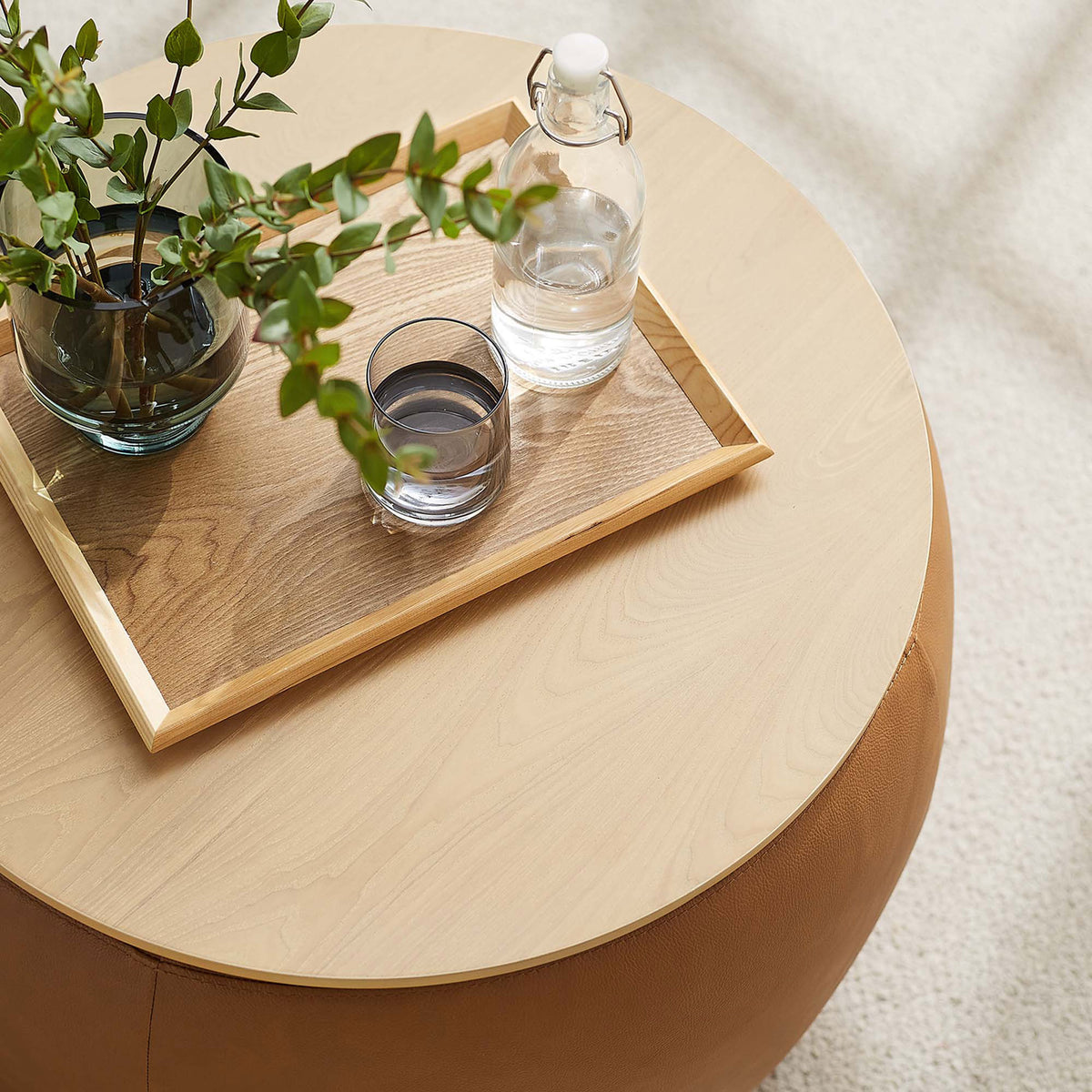 Tan round storage ottoman used as a coffee table with tray, glass, and greenery in a bright room || EEI-6687-TAN || front_angle_room