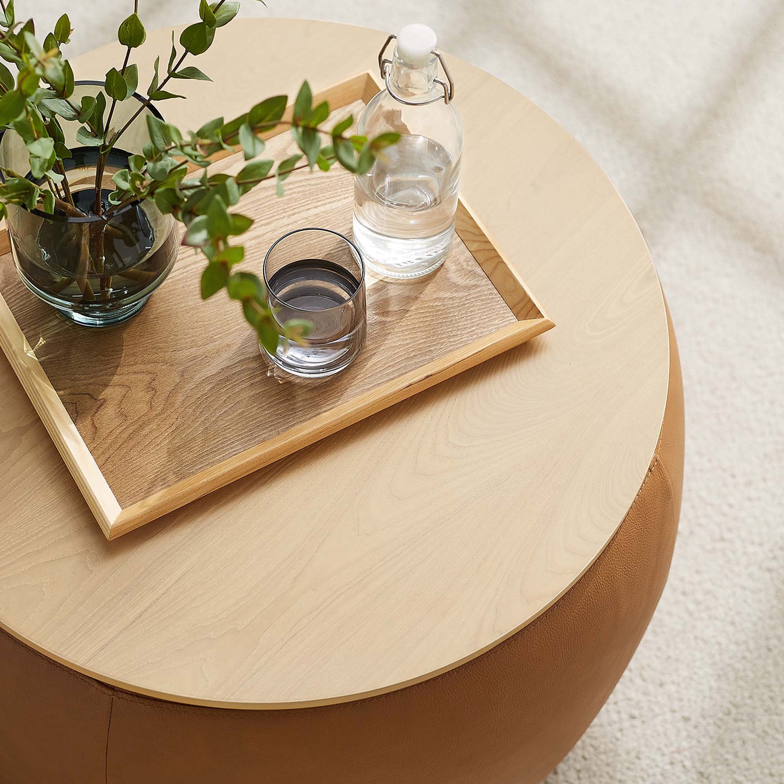 Tan round storage ottoman used as a coffee table with tray, glass, and greenery in a bright room || EEI-6687-TAN || front_angle_room