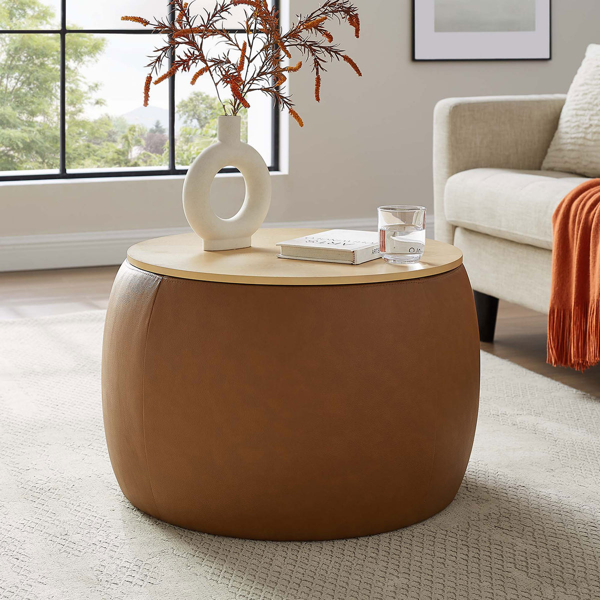 Tan round storage ottoman styled as a coffee table beside a modern sofa in a sunlit living room || EEI-6687-TAN || front_angle_room