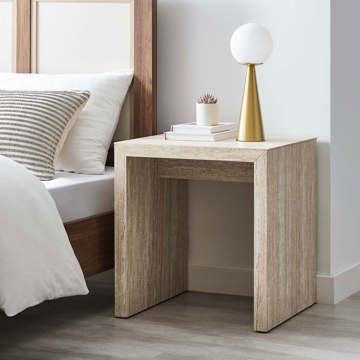Mirella faux travertine side table used as a nightstand beside the bed with a lamp and stacked books || EEI-6772-TRA || front_angle_room