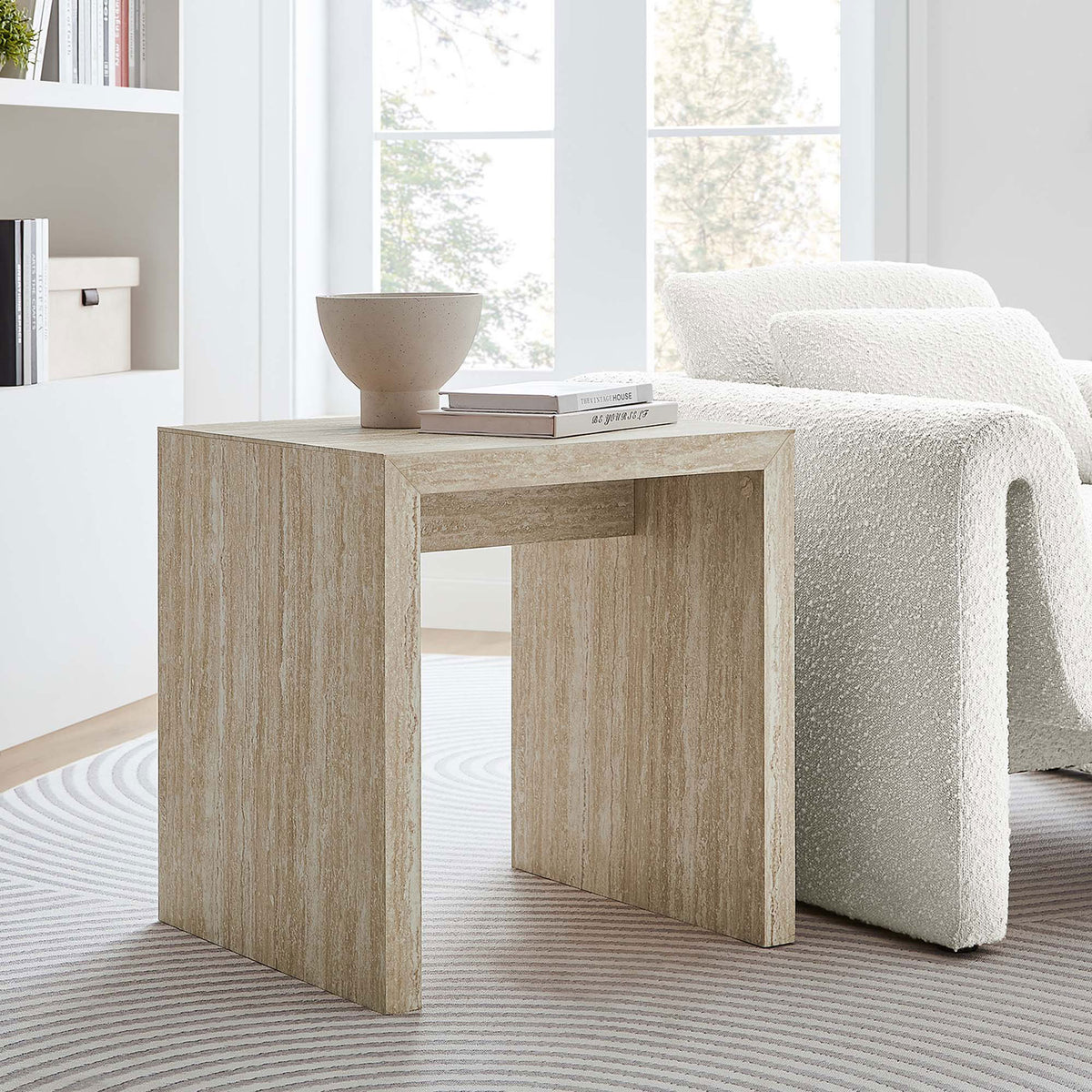 Mirella faux travertine side table paired with a curved sofa and round rug for a bright, modern living room || EEI-6772-TRA || front_angle_room