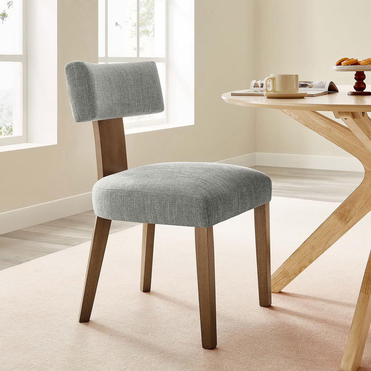 Upholstered dining chair paired with a round wood table in a bright, airy dining room. || EEI-6777-HEW-DBR || front_angle_partial_room