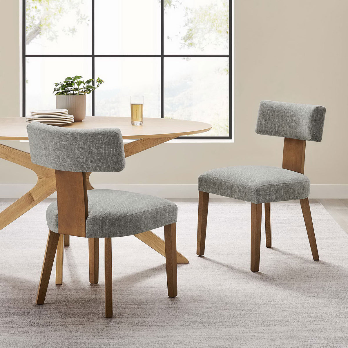 Pair of upholstered dining chairs surrounding a modern wood dining table in a sunlit room. || EEI-6777-HEW-DBR || front_angle_multi_room