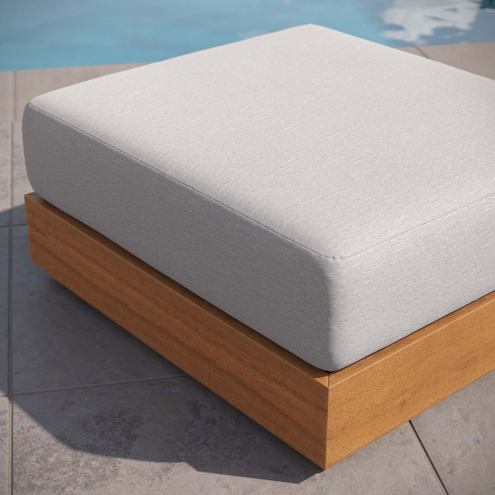 Tahoe ottoman with light gray cushion rests on a stone patio beside a shimmering swimming pool || EEI-6785-NAT-LGR || front_angle_room