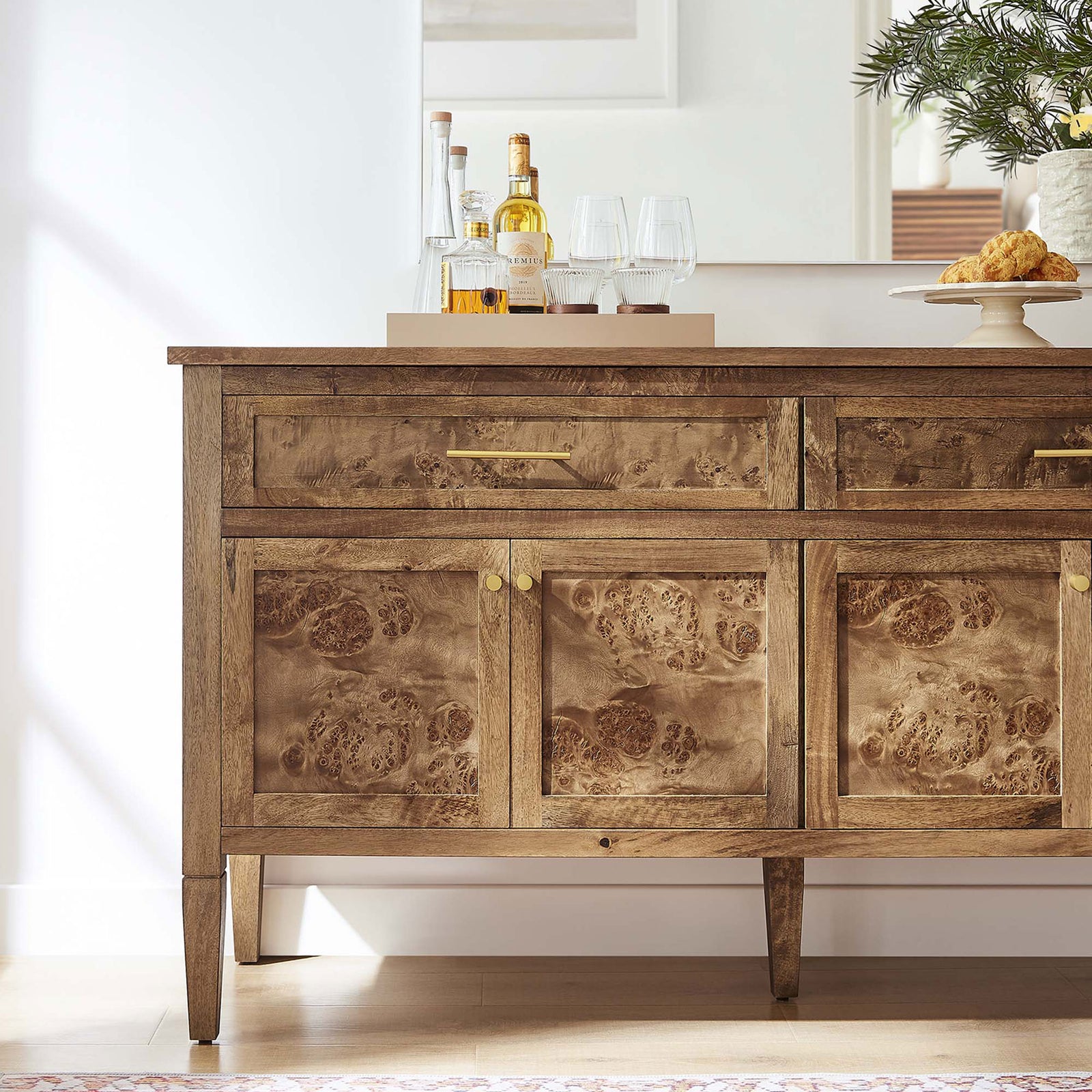 Styled burl wood sideboard used as a bar cabinet with trays, bottles, and glassware in a bright dining room. || EEI-6794-BRN || detail_room