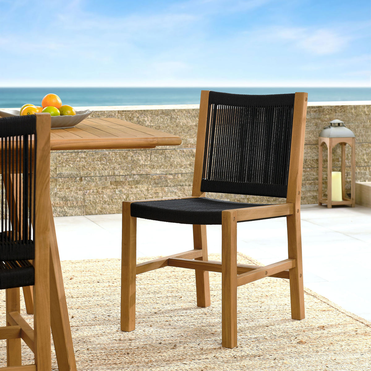 Teak and black rope dining chair styled by a coastal table setting on a sunlit outdoor terrace || EEI-6805-NAT-BLK || front_angle_room