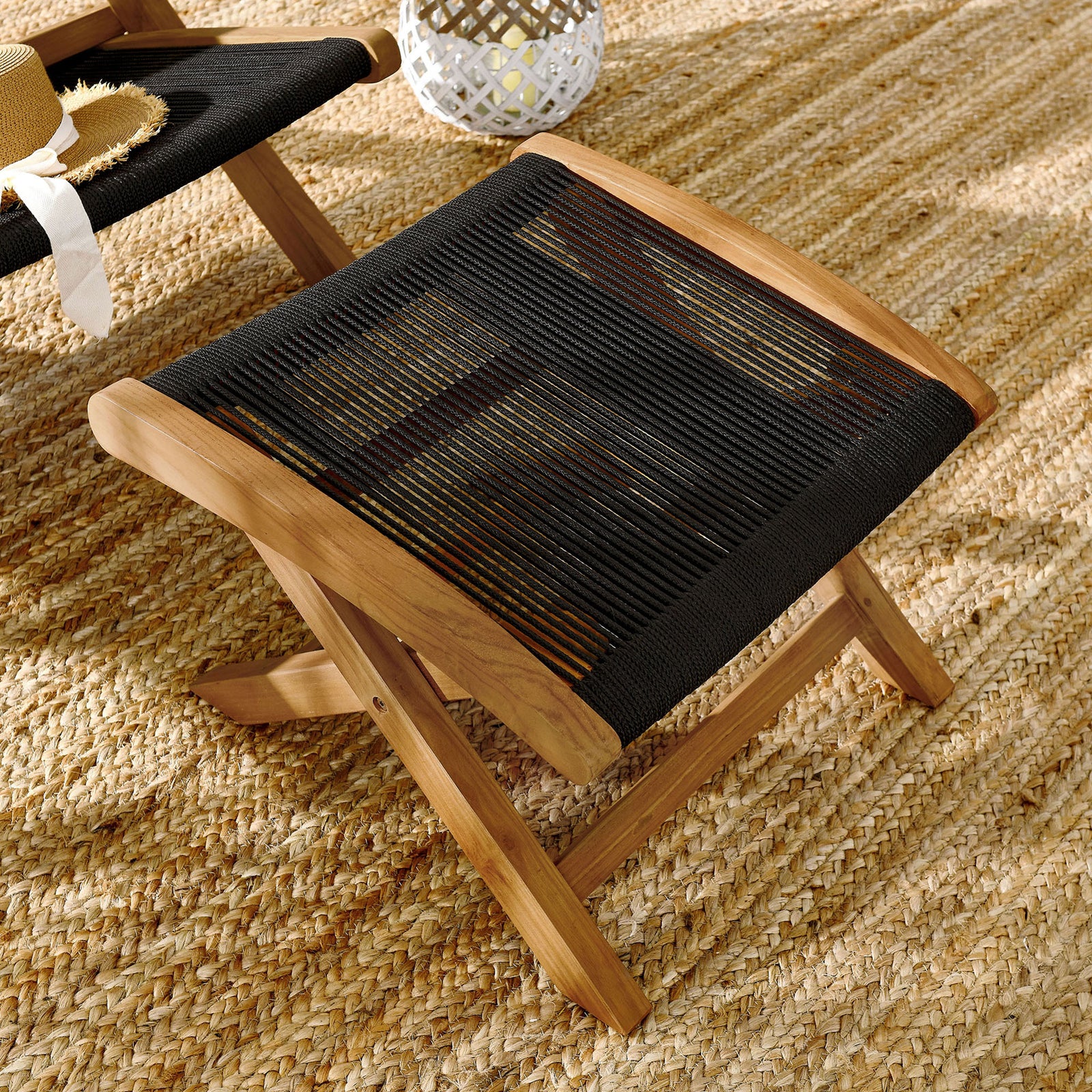 Teak folding ottoman with black rope seat styled on a natural jute rug for outdoor lounging || EEI-6809-NAT-BLK || front_angle_room