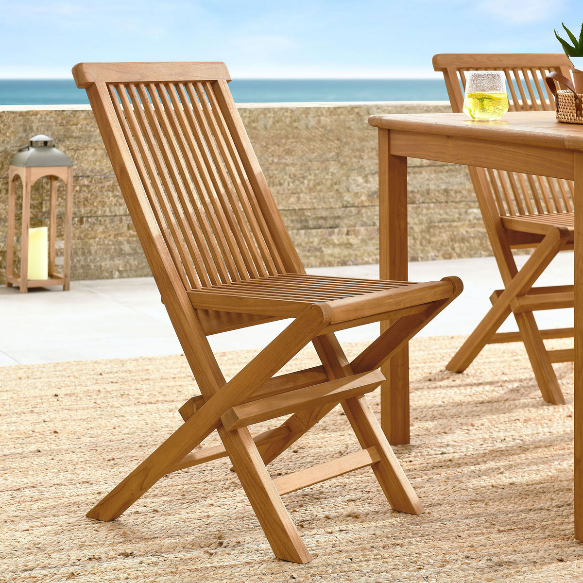 Natural teak folding dining chair styled on a coastal patio beside a matching outdoor dining table || EEI-6812-NAT || front_angle_room