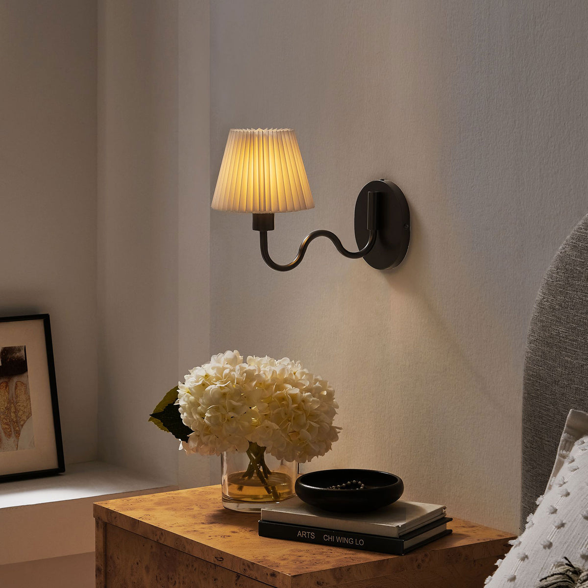 Glowing black squiggle arm wall sconce with pleated shade creates a cozy bedside reading light above the nightstand. || EEI-6873-BLK || front_angle_on_room