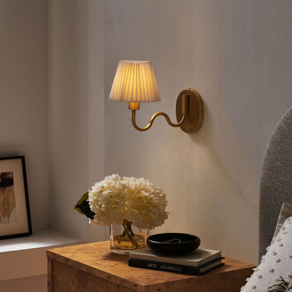 Illuminated brass squiggle arm wall sconce creates a cozy bedside glow above a nightstand styled with flowers. || EEI-6873-BRA || front_angle_on_room