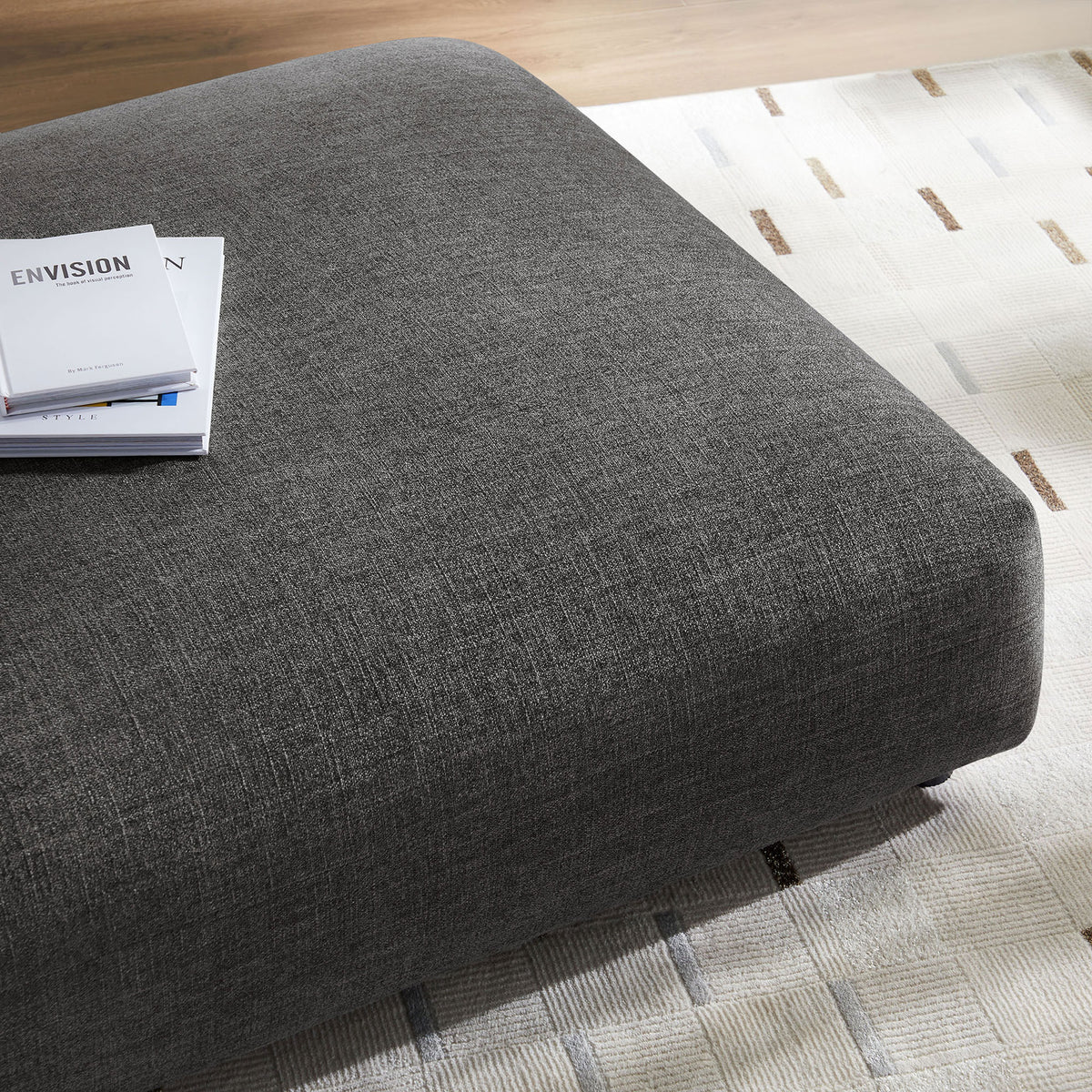 Charcoal ottoman styled on a patterned area rug with books to showcase its spacious, cushioned surface || EEI-6889-CHA || detail_room