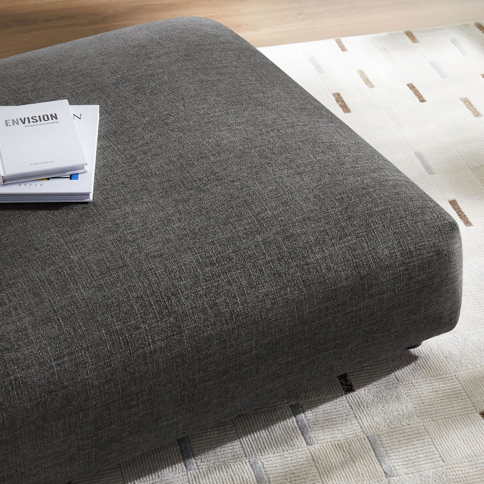 Charcoal ottoman styled on a patterned area rug with books to showcase its spacious, cushioned surface || EEI-6889-CHA || detail_room