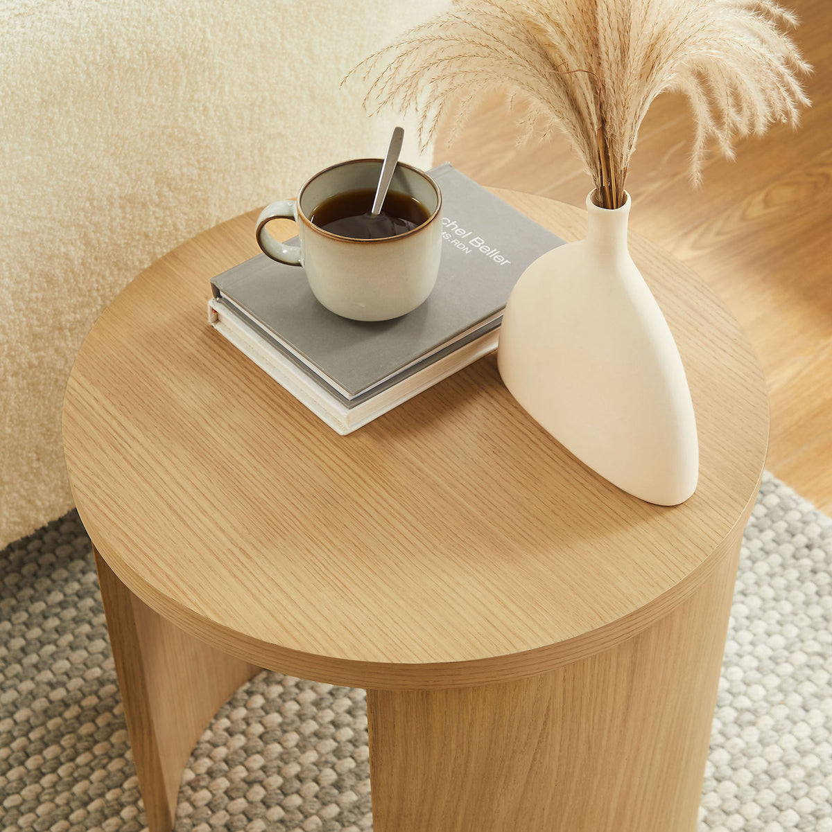 Oak round end table styled with a mug, book, and vase on a textured neutral rug || EEI-6913-OAK || front_angle_room