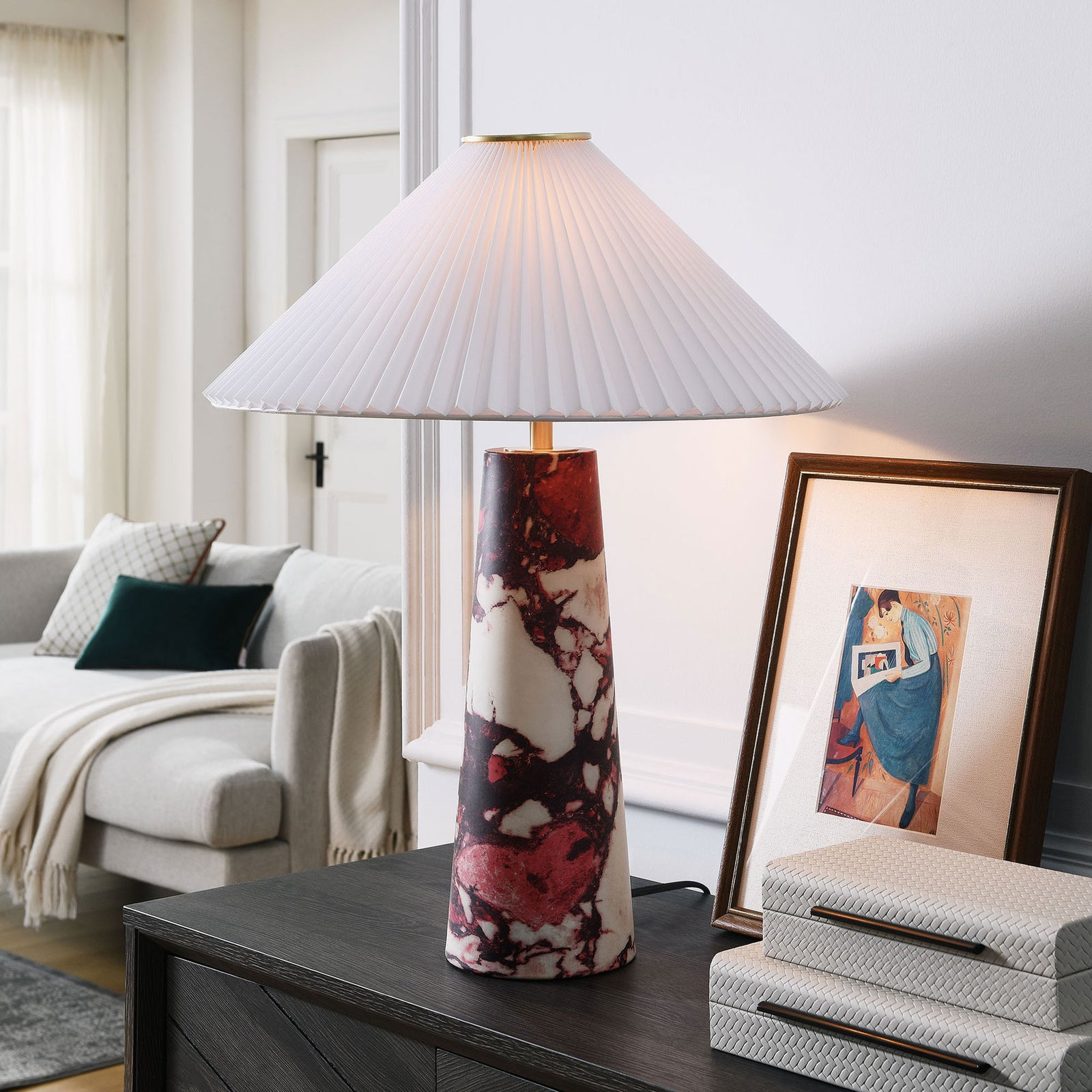 Red marble Nera table lamp glowing beside framed art and stacked boxes on a console. || EEI-6963-RED || front_on_room