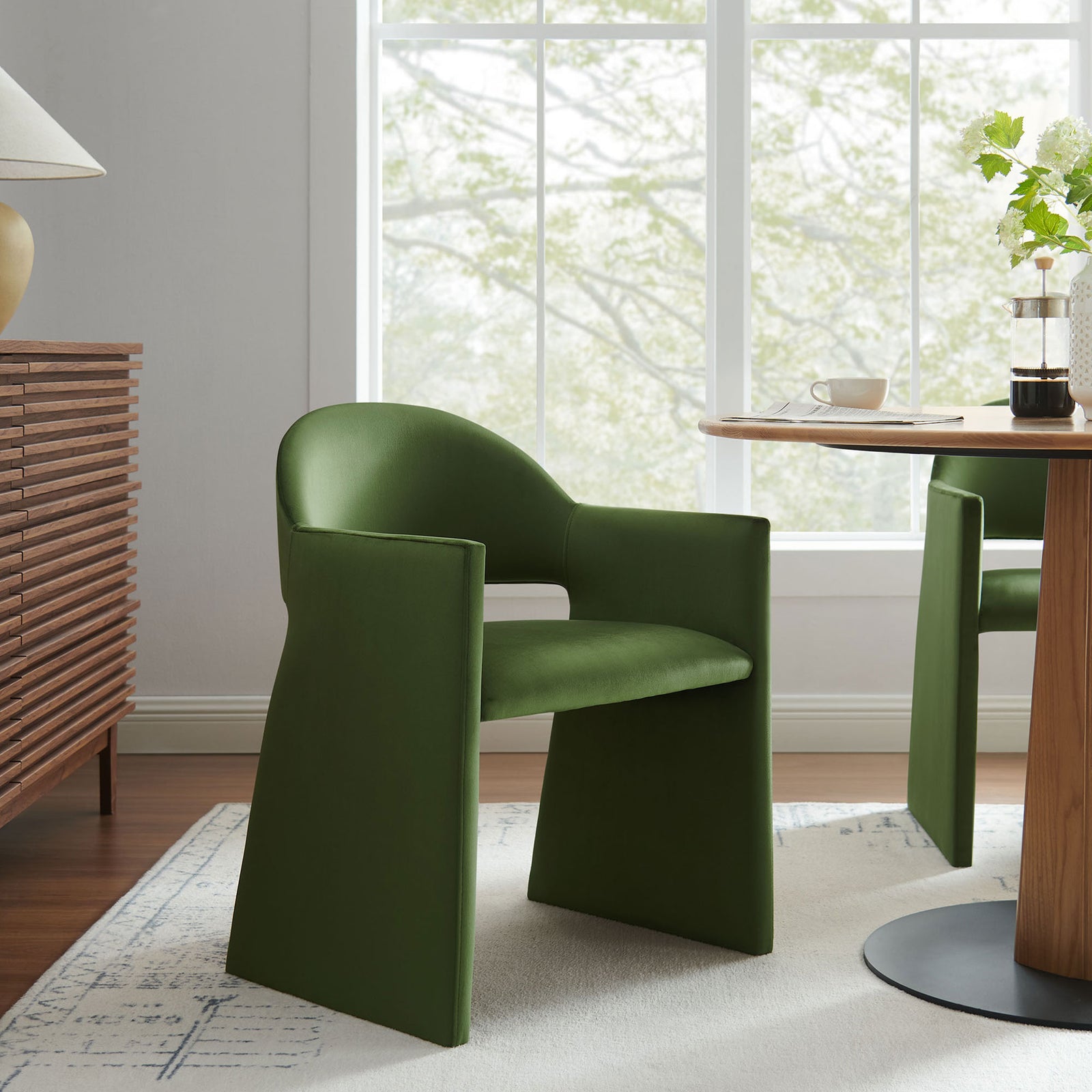 Moss green velvet dining armchair styled at a modern round table, bringing sculptural comfort to a bright dining room. || EEI-7081-MOG || front_angle_room