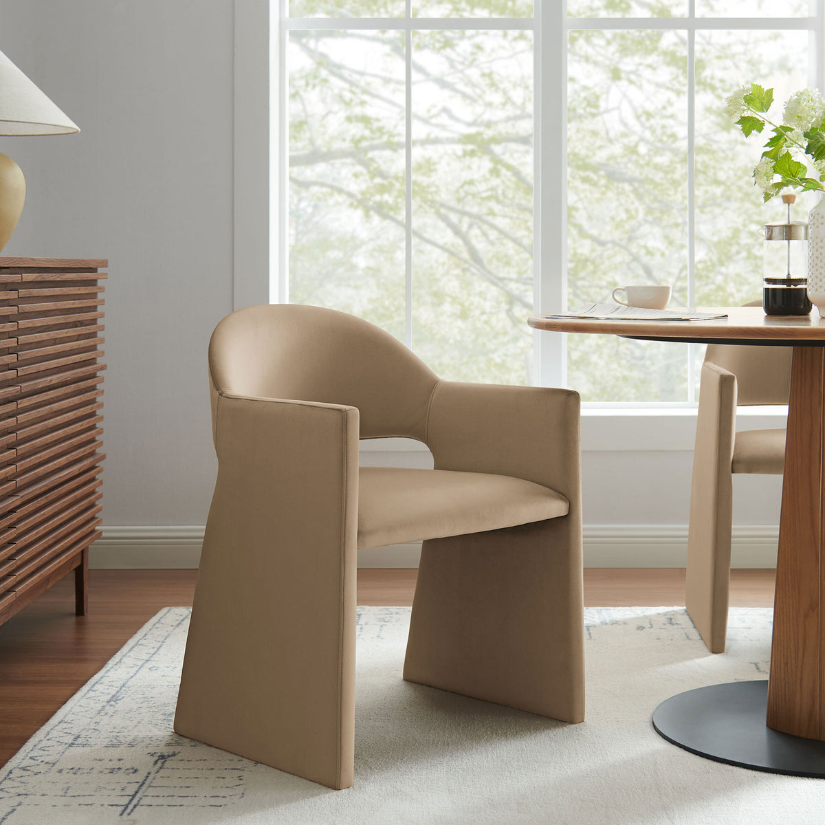 Taupe velvet dining armchair styled in a bright dining room beside a round wood table and sideboard || EEI-7081-TAU || front_angle_room