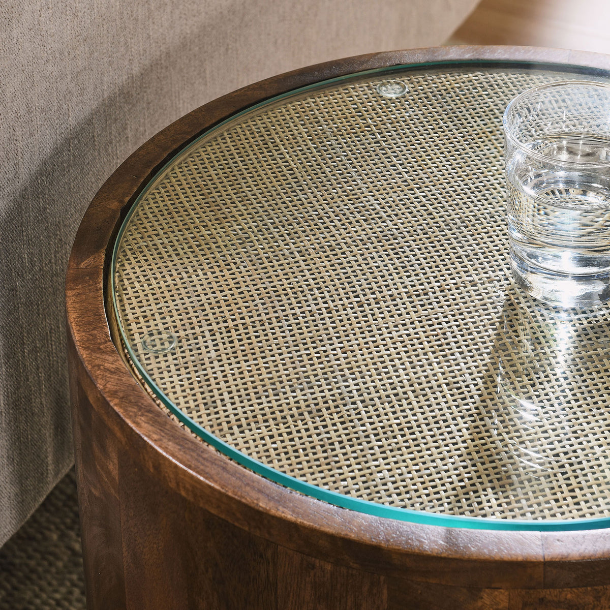 Glass-topped rattan cane side table holding a drink beside a sofa in a cozy living room || EEI-7085-WAL || detail_room