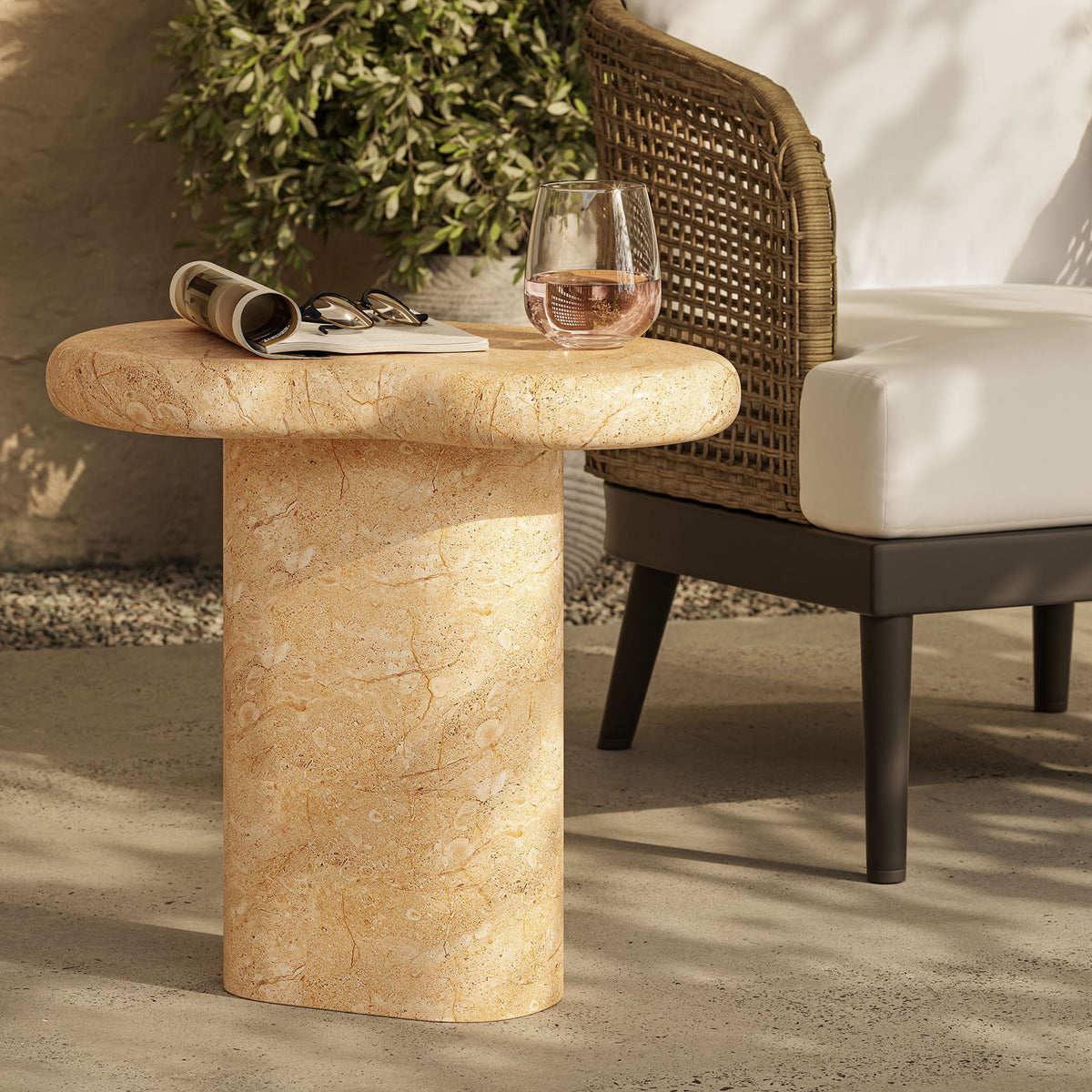 Textured travertine-look side table on a sunlit patio beside woven lounge seating, styled with a drink and book || EEI-7094-TRV || front_angle_room