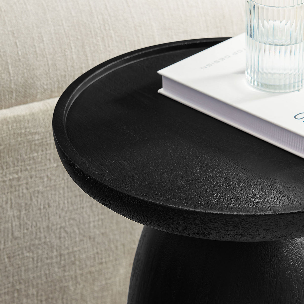 Black tray-top side table styled with a glass and book beside a light, textured sofa. || EEI-7145-BLK || detail_room
