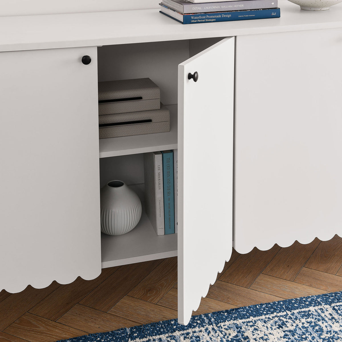 White scalloped sideboard styled with books and decor, showcasing interior shelving on a blue patterned rug || EEI-7153-WHI || front_open_room