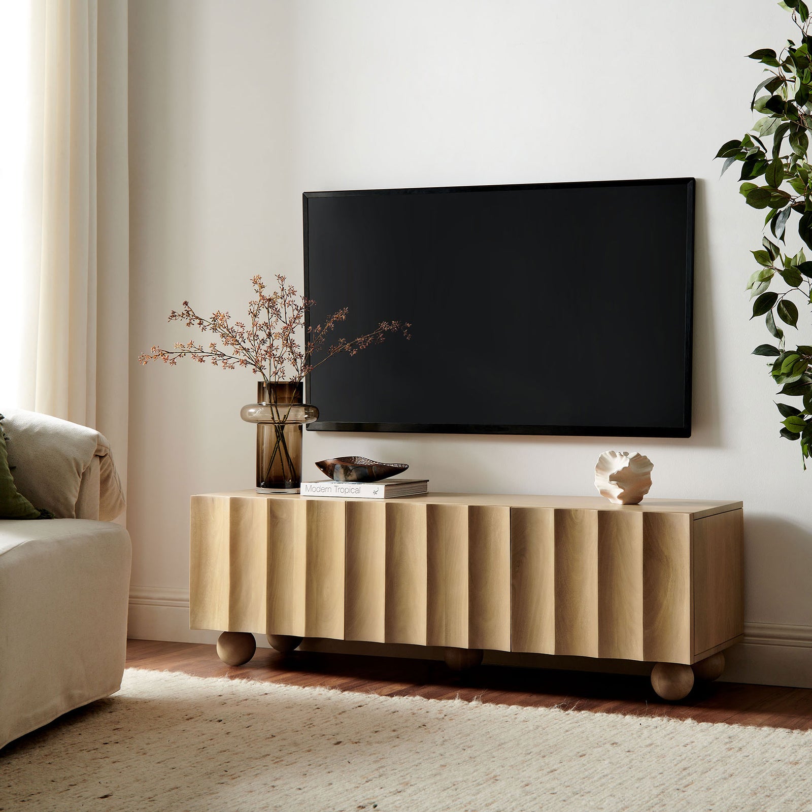 Natural wood TV stand styled beneath a wall-mounted TV with decor accents and rounded bun feet || EEI-7183-NAT || front_angle_room