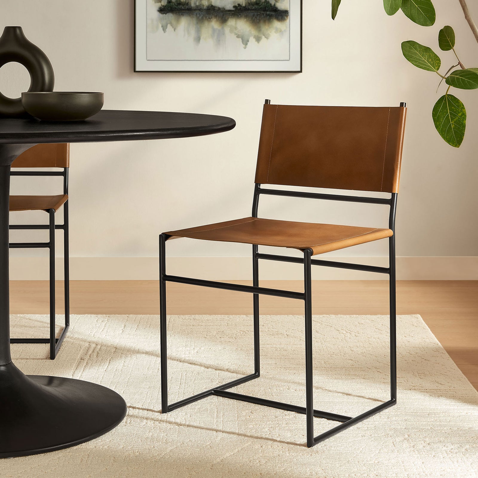 Tan leather and black metal sled dining chair styled in a modern room beside a pedestal table || EEI-7196-TAN || front_angle_room