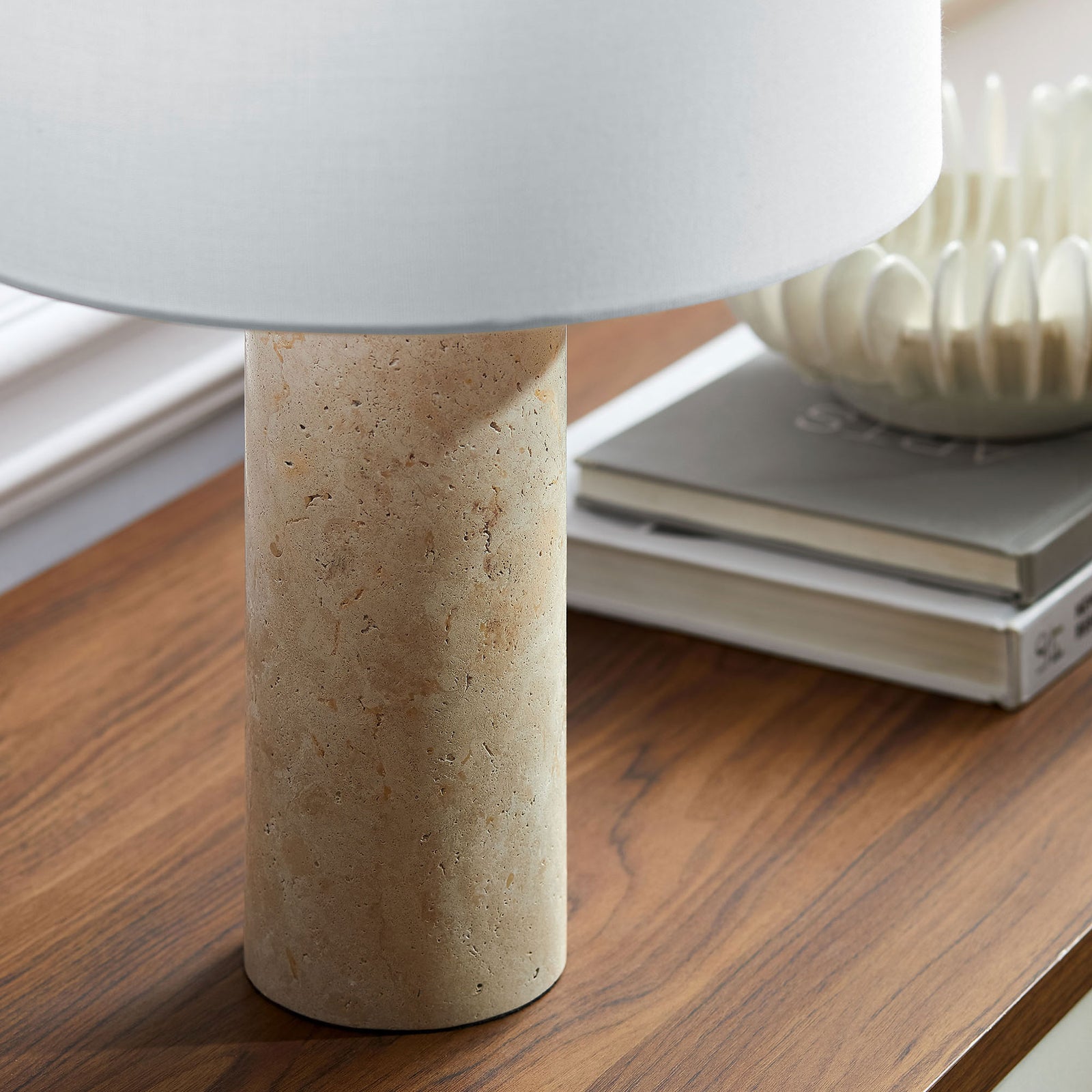 Travertine lamp base resting on a wood console layered with books and decorative bowl || EEI-7213-TRV || detail_room