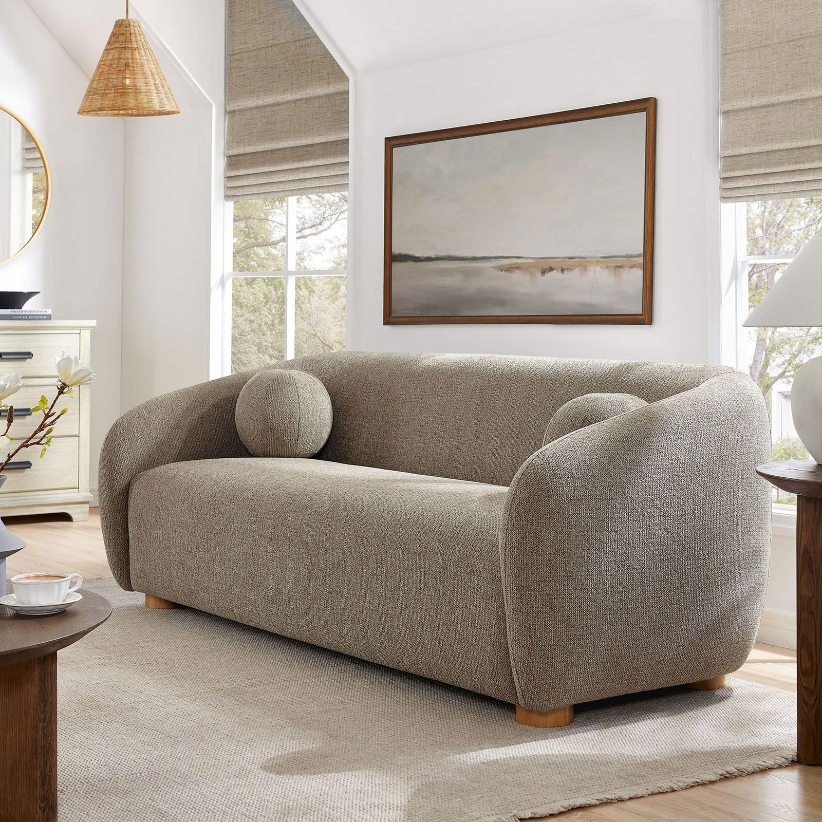 Taupe curved sofa with ball pillows styled in a cozy living room with natural wood accents and soft rug || EEI-7217-TAU || front_angle_room