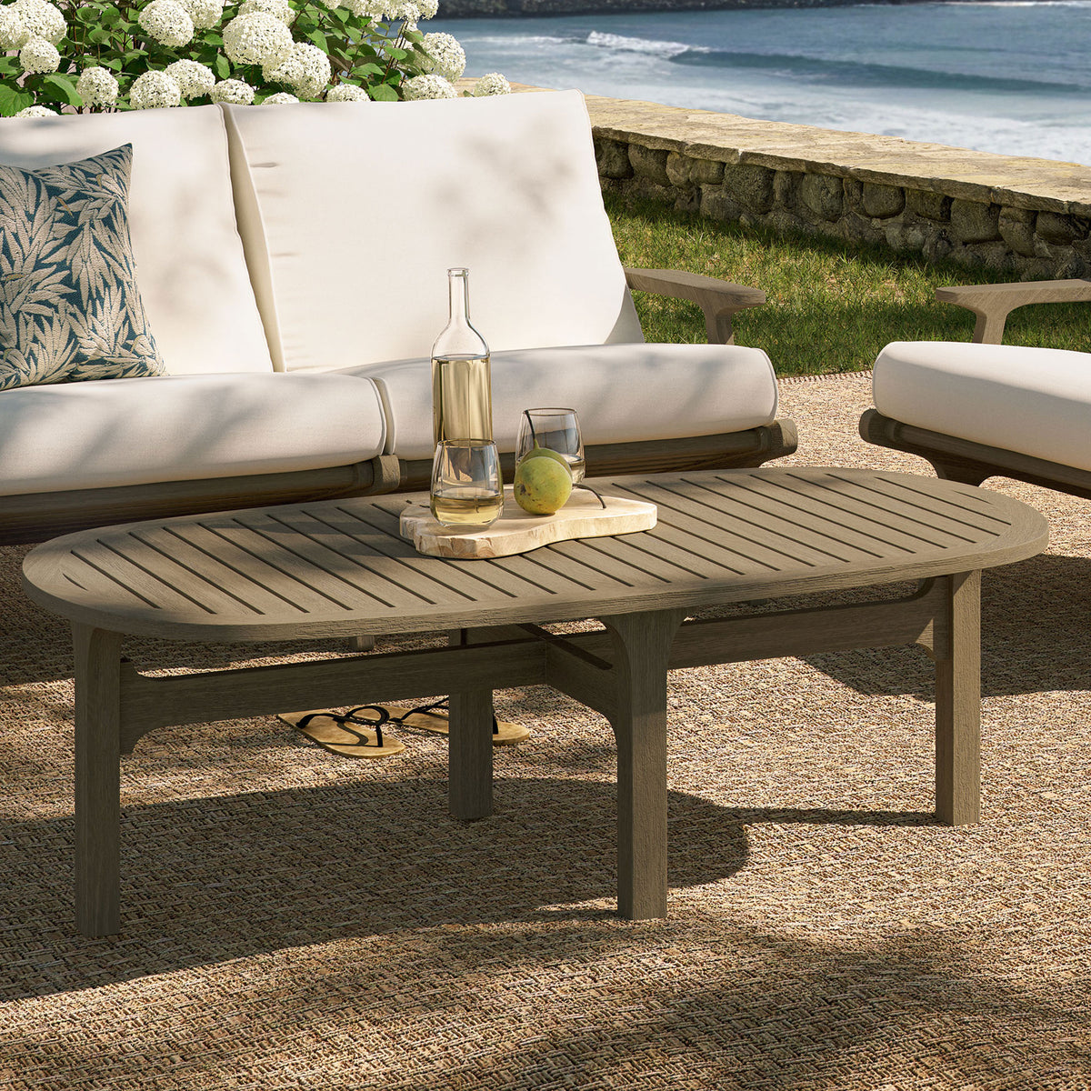 Saratoga oval outdoor teak coffee table in weathered gray styled on a coastal patio seating area || EEI-7246-WGR || front_angle_room