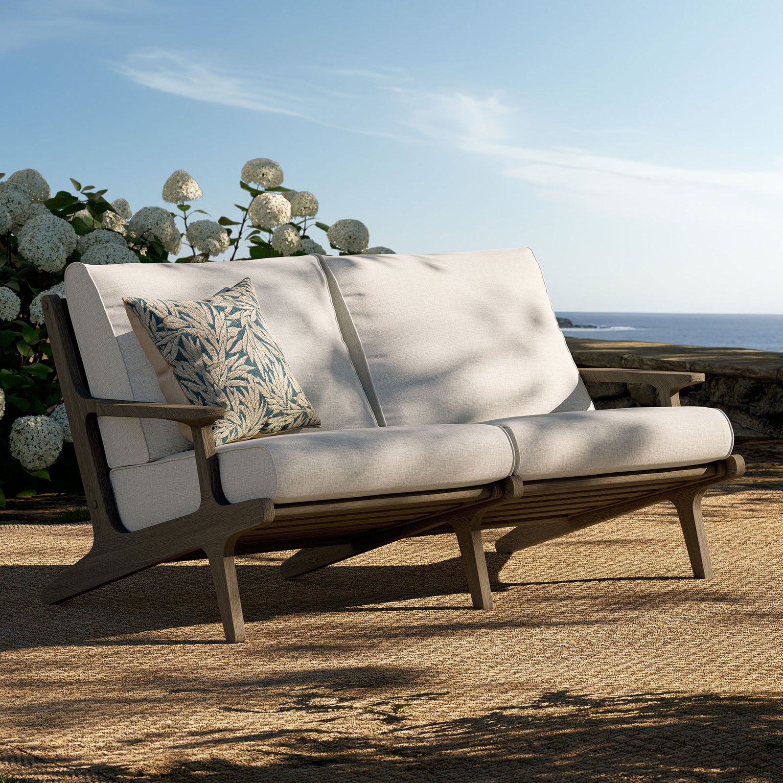 Saratoga teak loveseat styled on a sunlit patio with plush pebble cushions and decorative pillow. || EEI-7247-WGR-PEB || front_angle_room