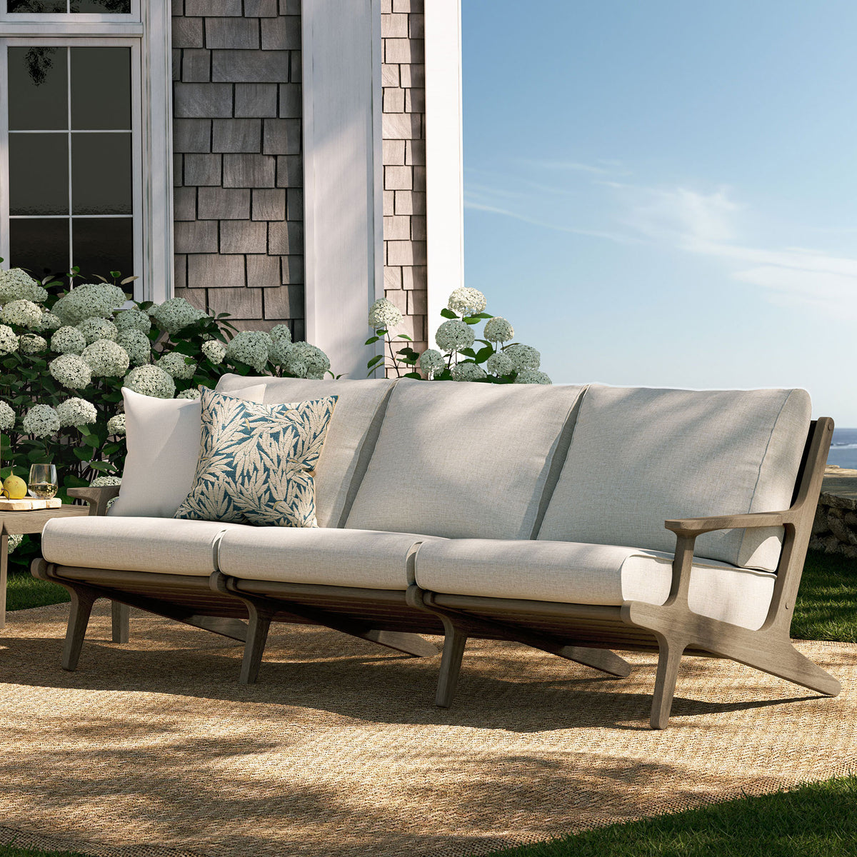 Saratoga teak sofa on a sunlit patio, styled with pebble cushions and botanical accent pillows || EEI-7249-WGR-PEB || front_angle_room