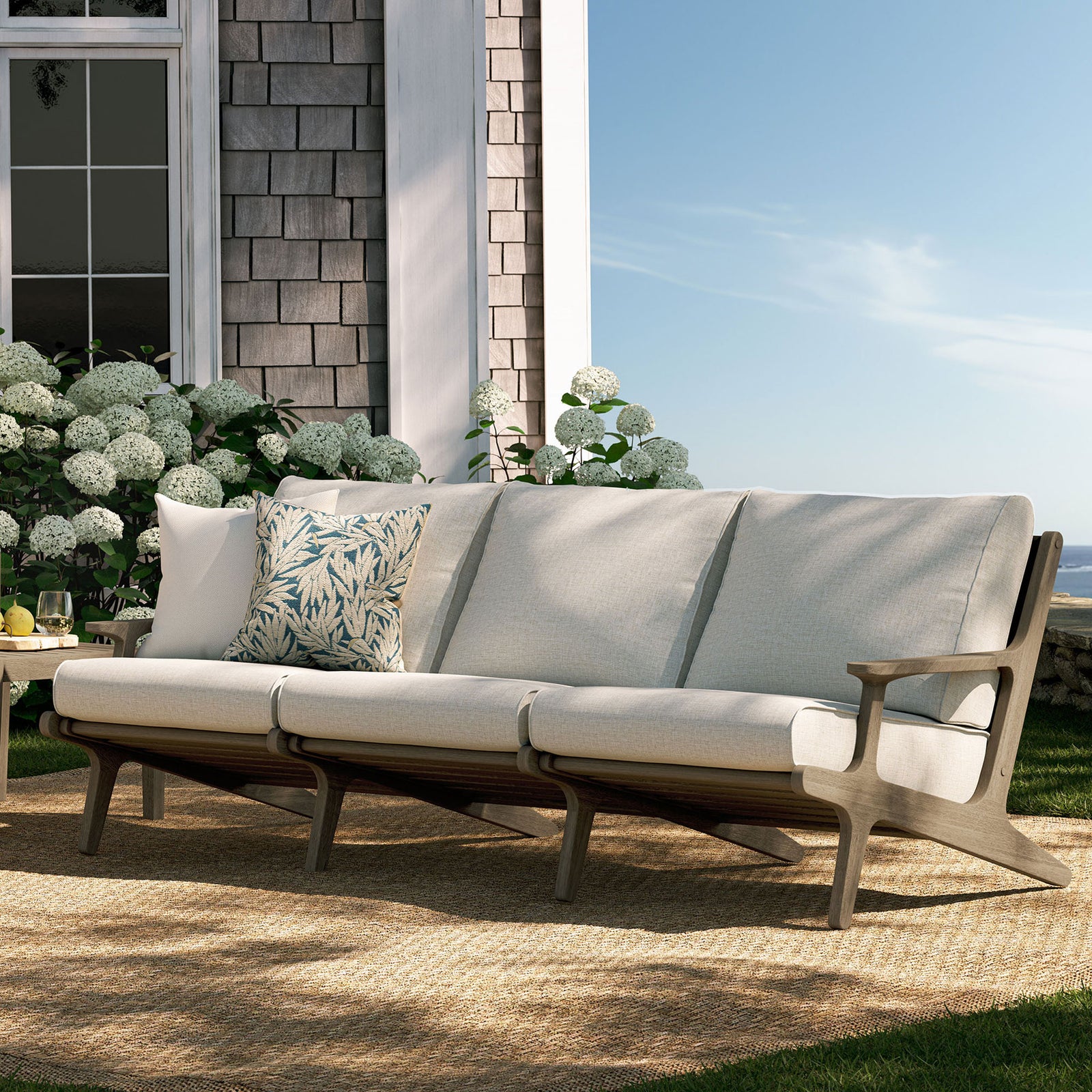 Saratoga teak sofa on a sunlit patio, styled with pebble cushions and botanical accent pillows || EEI-7249-WGR-PEB || front_angle_room