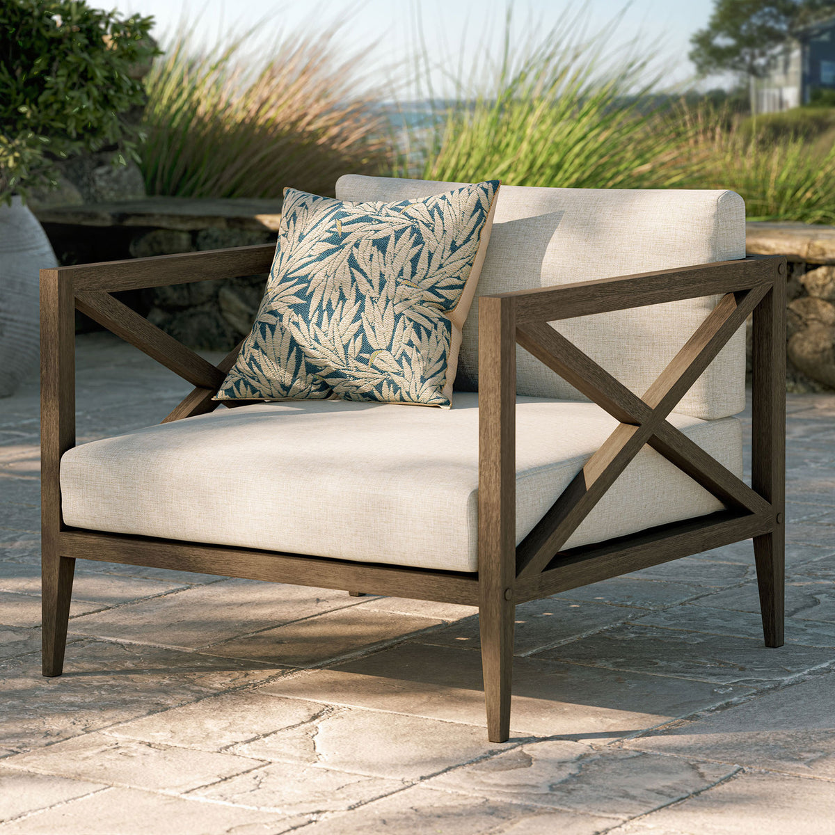 Northlake teak armchair in pebble fabric styled with a patterned pillow on a stone terrace setting || EEI-7252-WGR-PEB || front_angle_room