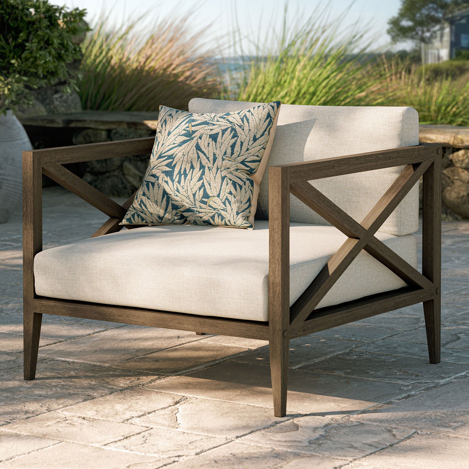Northlake teak armchair in pebble fabric styled with a patterned pillow on a stone terrace setting || EEI-7252-WGR-PEB || front_angle_room