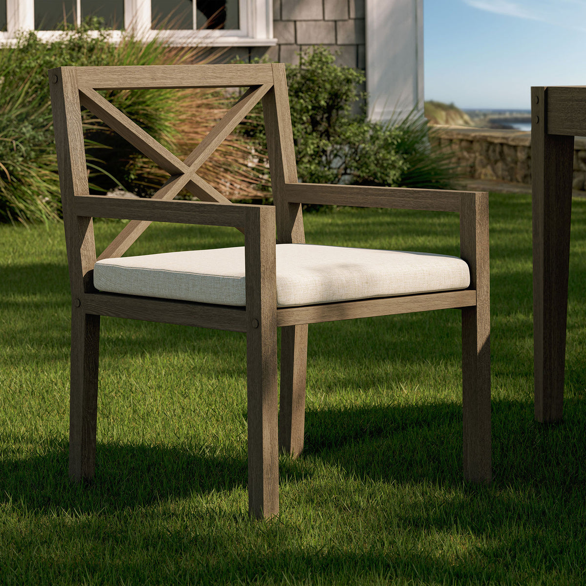 Northlake teak armchair with pebble cushion arranged on a green lawn beside an outdoor dining set. || EEI-7253-WGR-PEB || front_angle_room