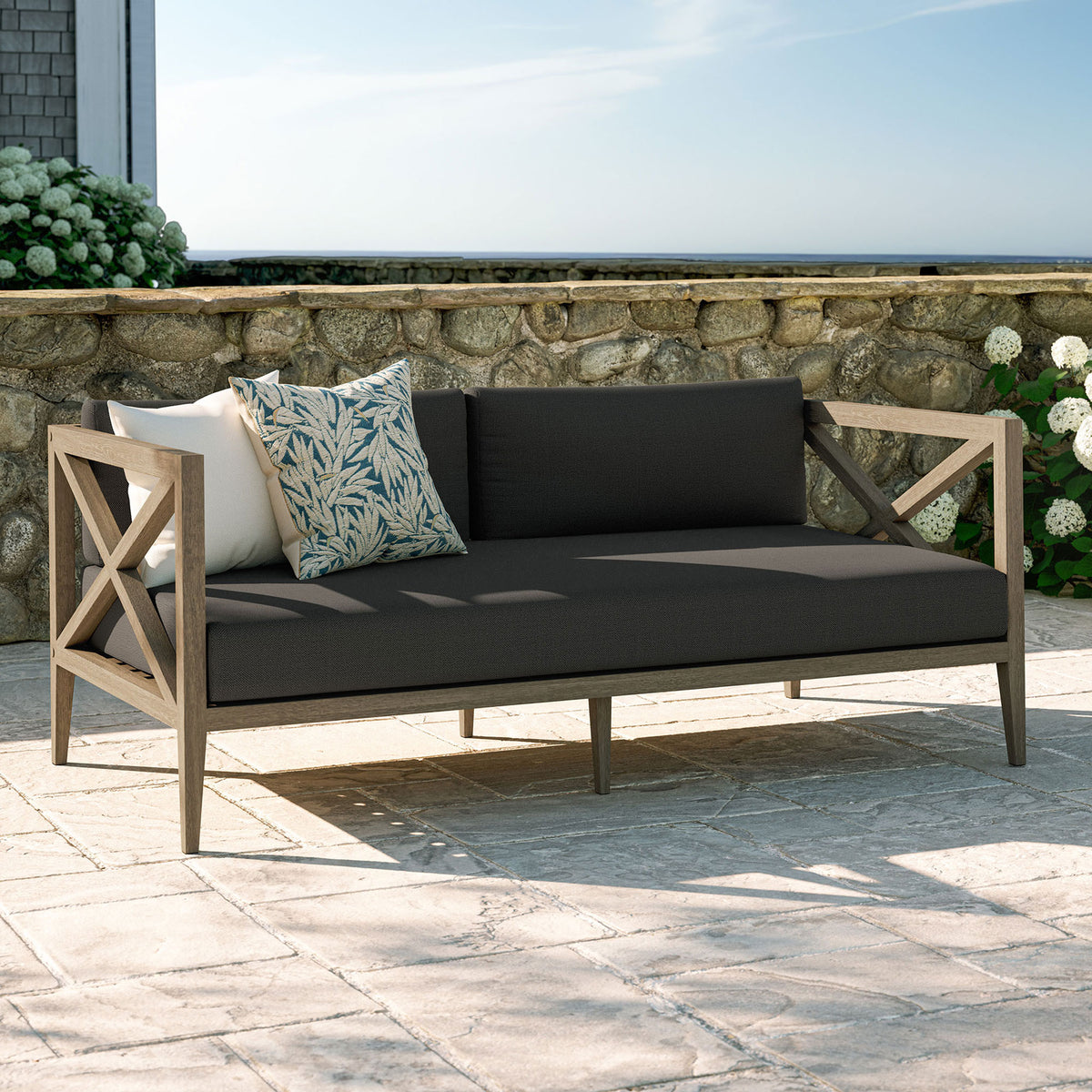 Flint gray cushioned teak loveseat styled on a stone patio, perfect for garden lounges and coastal decks. || EEI-7254-WGR-FLI || front_angle_room