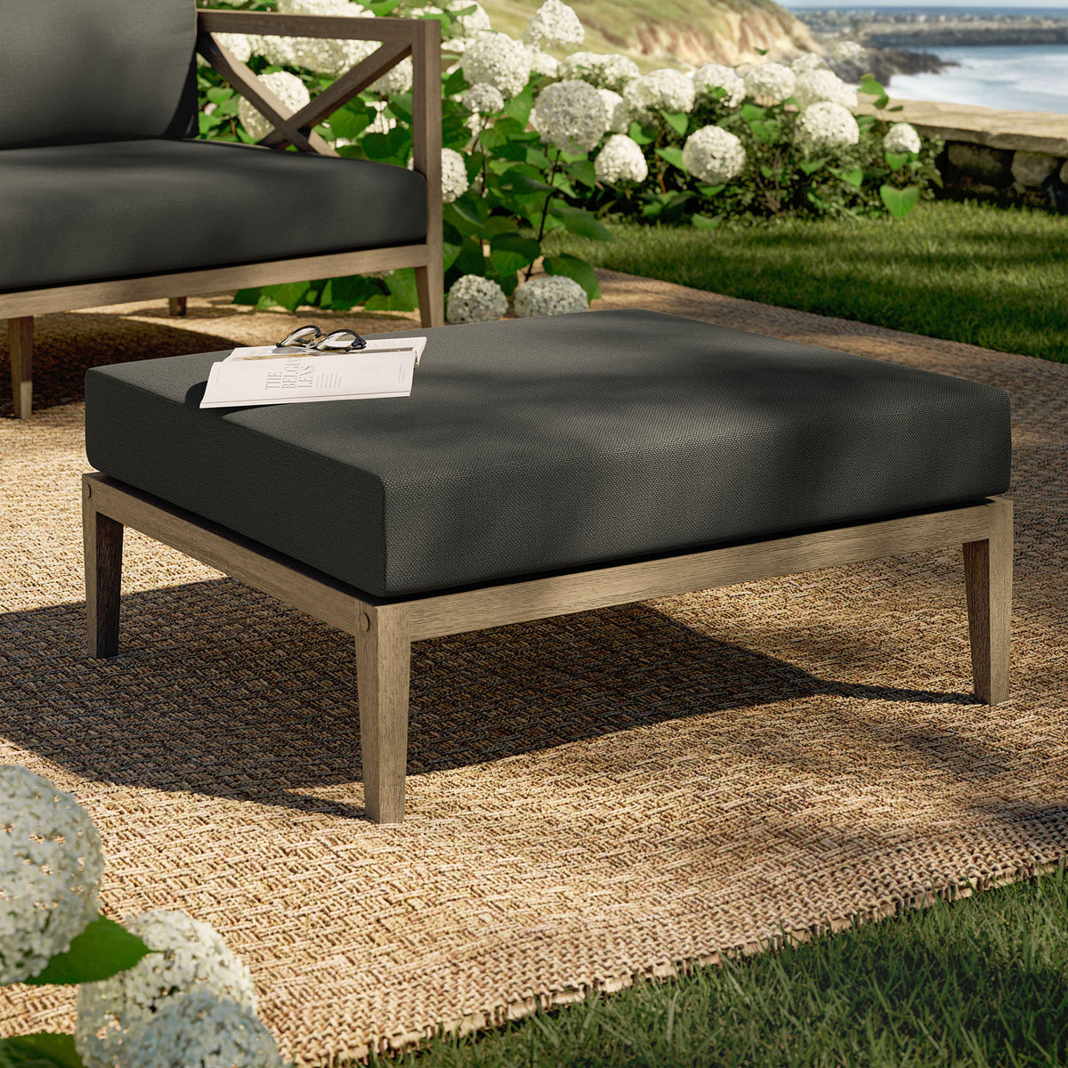 Teak patio ottoman with charcoal cushion styled on a woven rug amid lush garden greenery || EEI-7255-WGR-FLI || front_angle_room