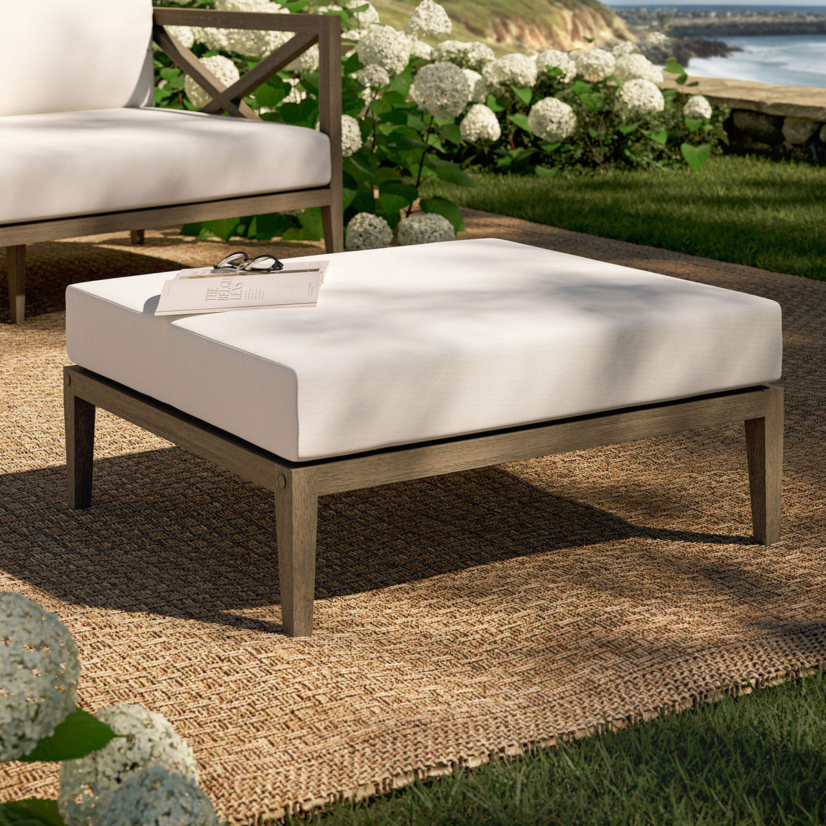 Teak patio ottoman with white cushion styled on a woven rug amid lush garden greenery || EEI-7255-WGR-WHI || front_angle_room