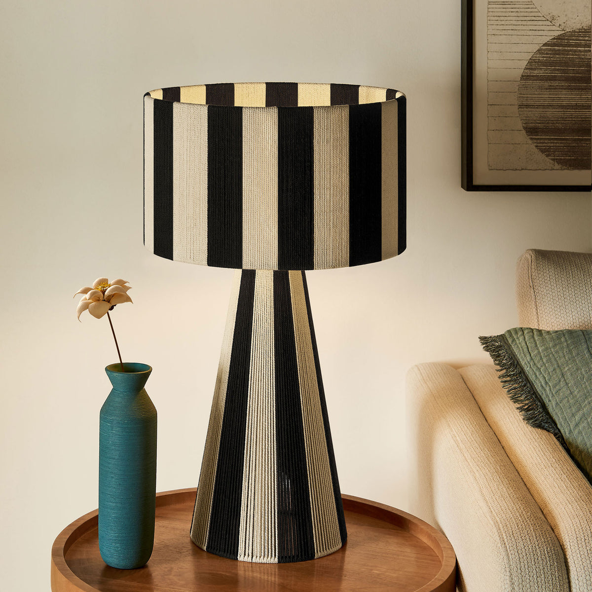 Illuminated black and cream striped table lamp casts a warm glow beside a cozy sofa. || EEI-7261-BLK || front_on_room