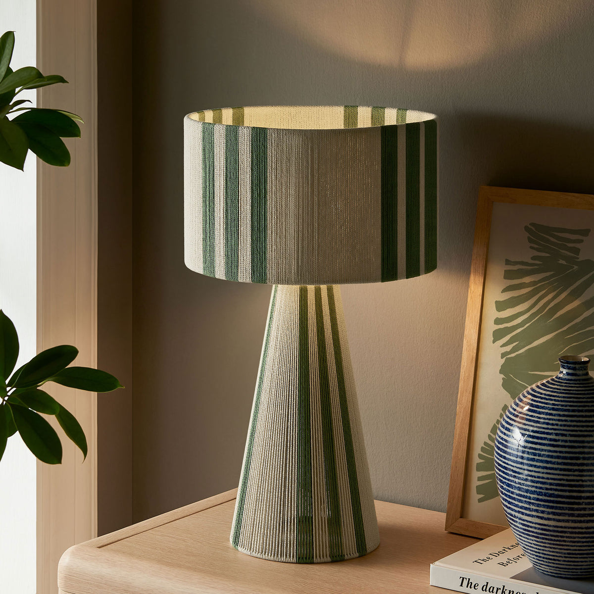 Basil green striped table lamp glowing softly to create a cozy, inviting atmosphere. || EEI-7262-BAS || front_angle_on_room
