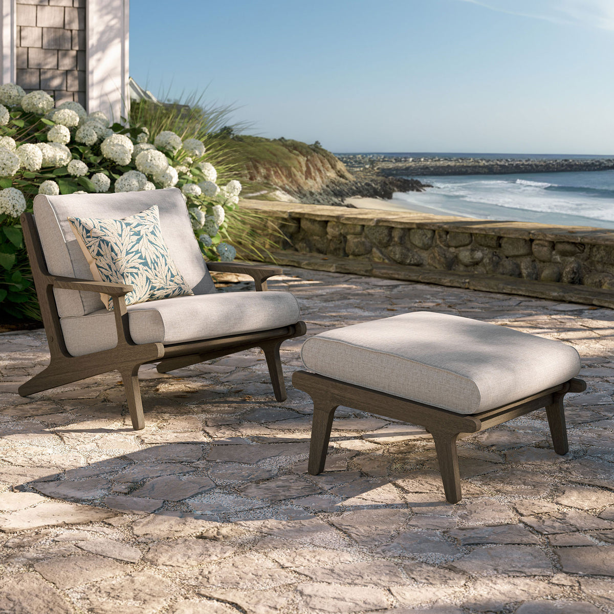 Saratoga pebble chair and ottoman styled on a sunny patio with ocean views and lush hydrangeas || EEI-7268-WGR-PEB || front_angle_room