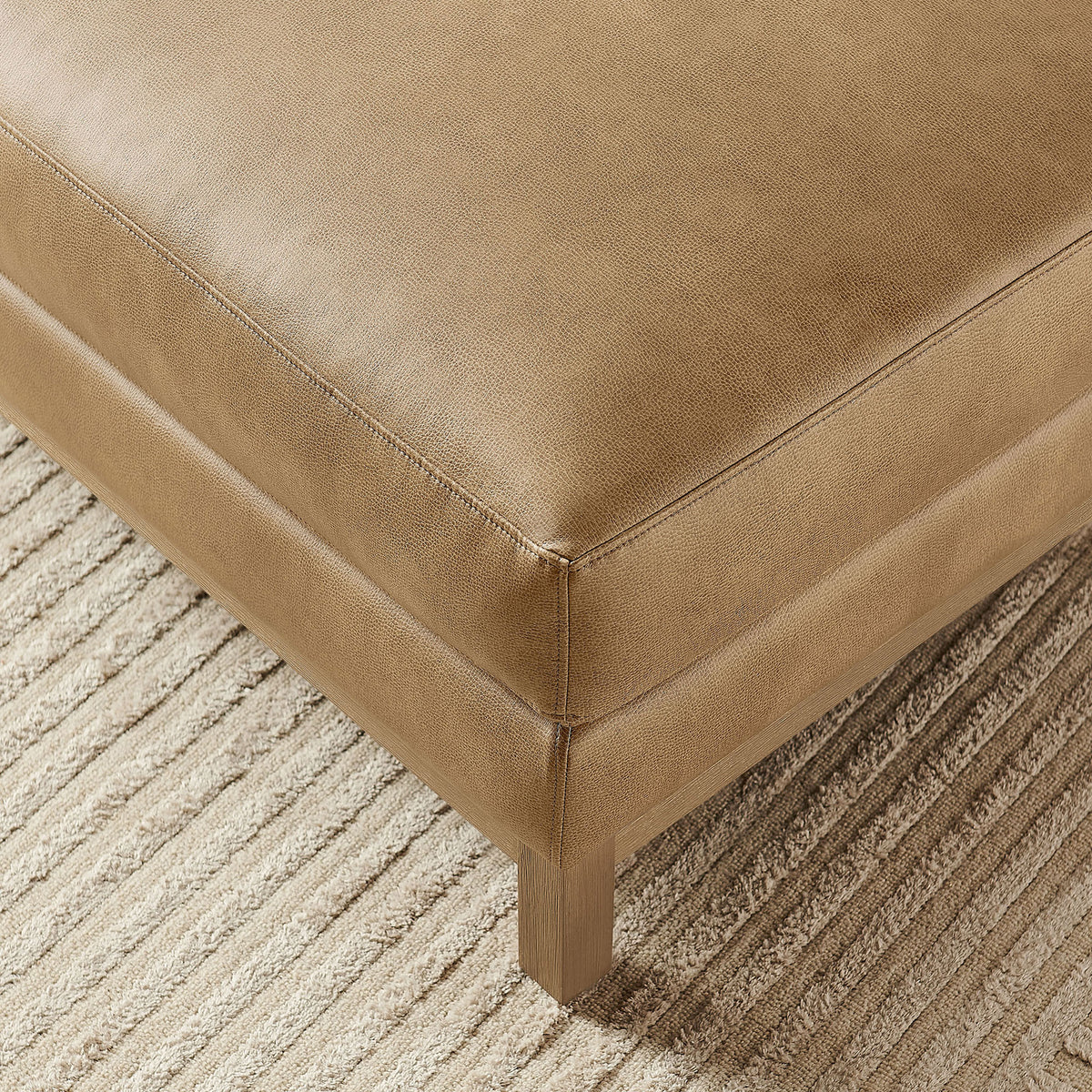 Corner of toast vegan leather ottoman resting on a textured area rug with warm tones || EEI-7338-TST || detail_room
