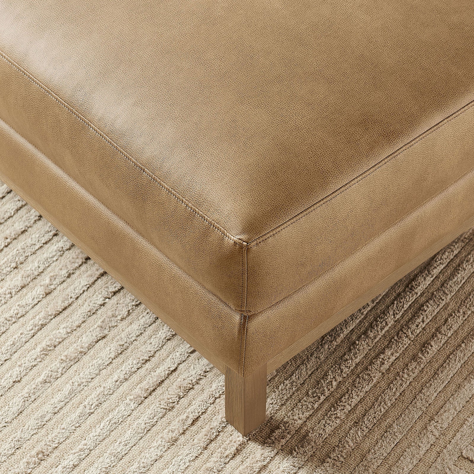 Corner of toast vegan leather ottoman resting on a textured area rug with warm tones || EEI-7338-TST || detail_room