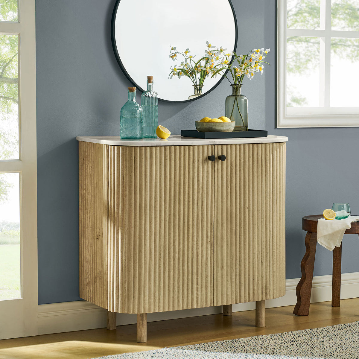 Curved fluted wood cabinet with marble top styled in a bright room with mirror, window, and decor accents || EEI-7354-NAT || front_room