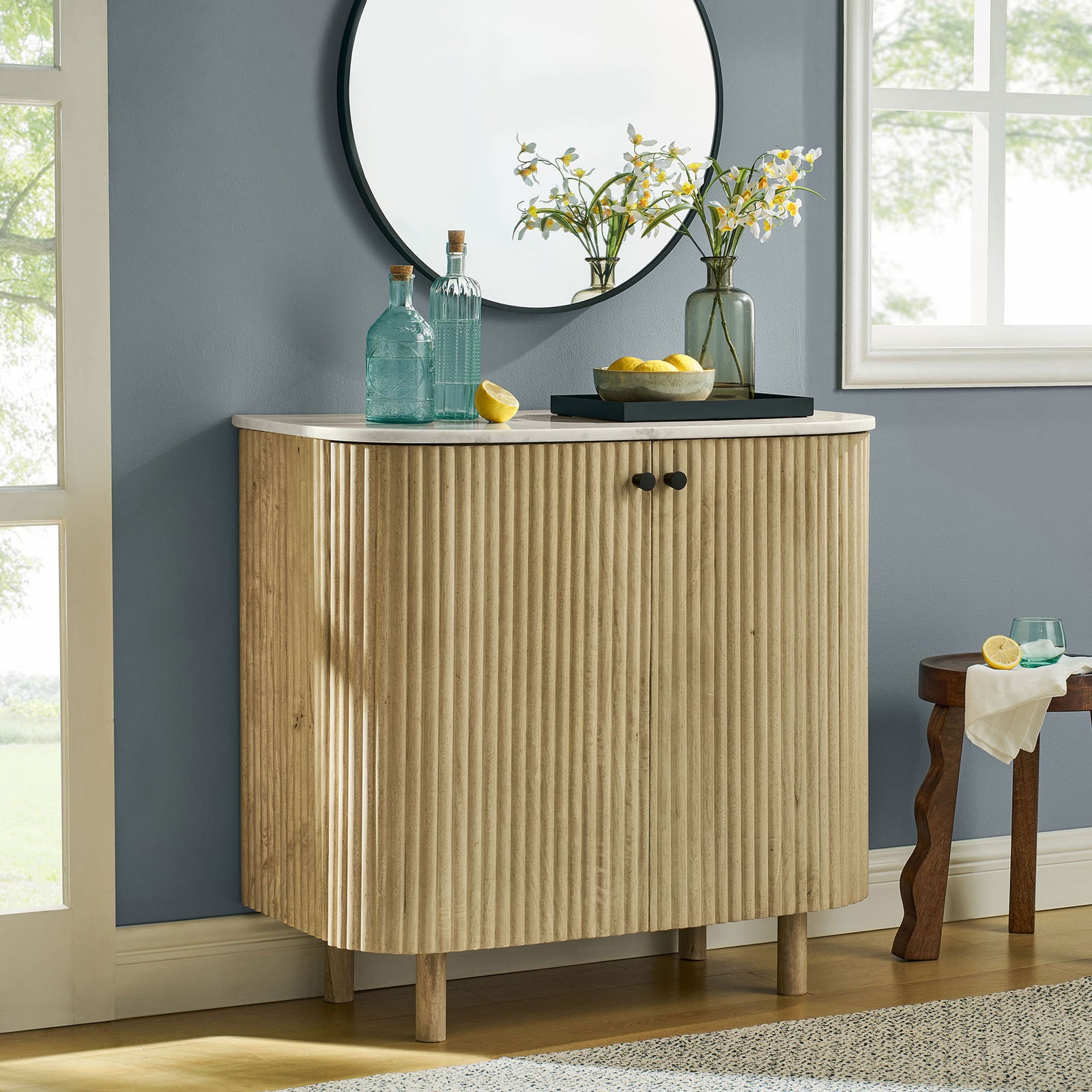 Curved fluted wood cabinet with marble top styled in a bright room with mirror, window, and decor accents || EEI-7354-NAT || front_room