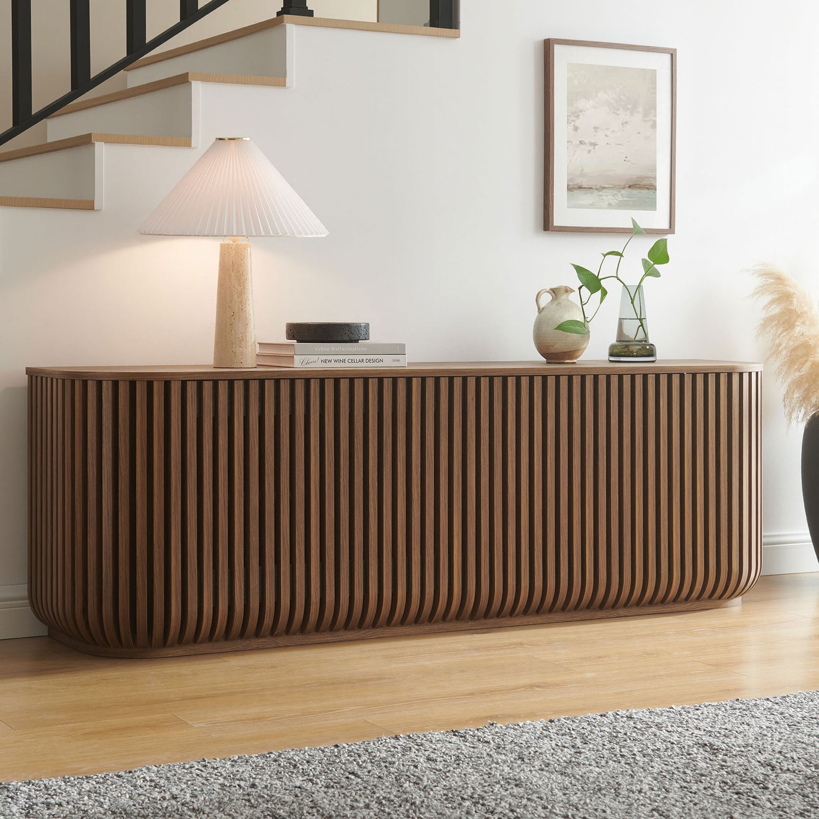 Curved walnut sideboard anchors a bright entry or living space, topped with sculptural decor and a table lamp || EEI-7359-WAL || front_angle_room