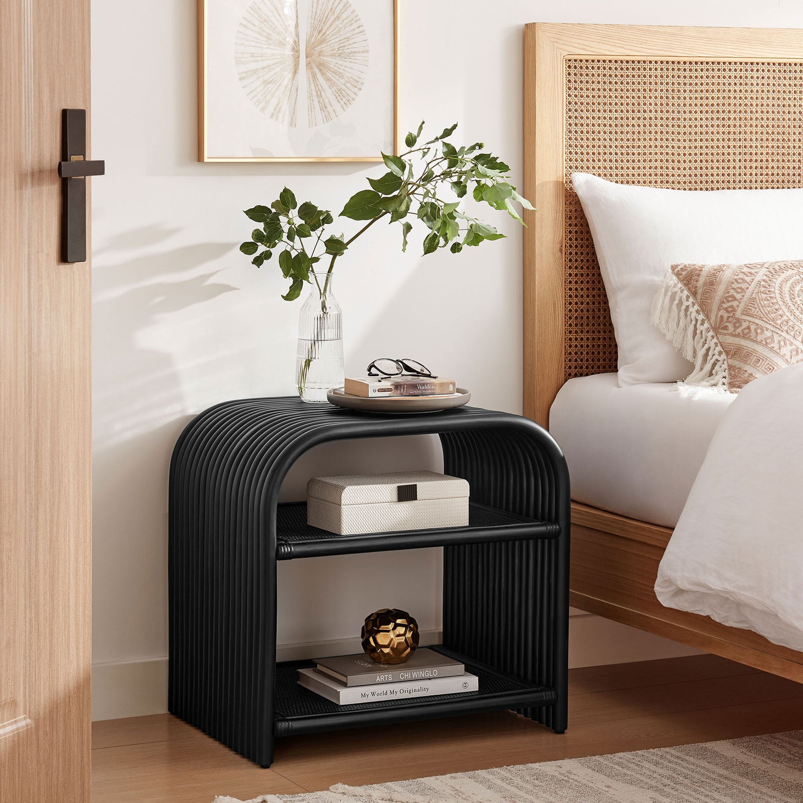 Black curved rattan side table used as a modern nightstand beside a warm neutral bed || EEI-7361-BLK || front_angle_room