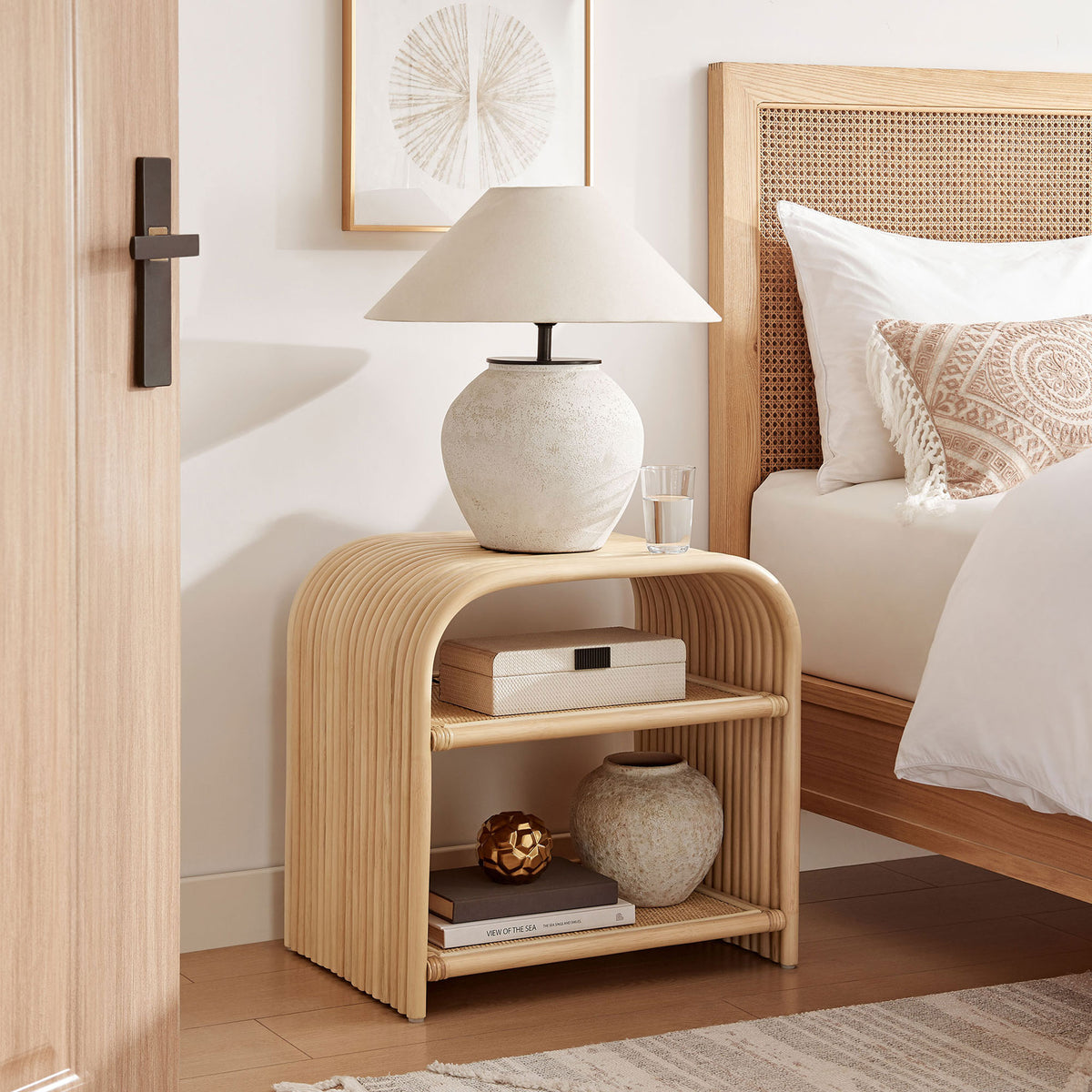 Curved natural rattan side table nightstand with woven shelves styled beside a neutral bed || EEI-7361-NAT || front_angle_room