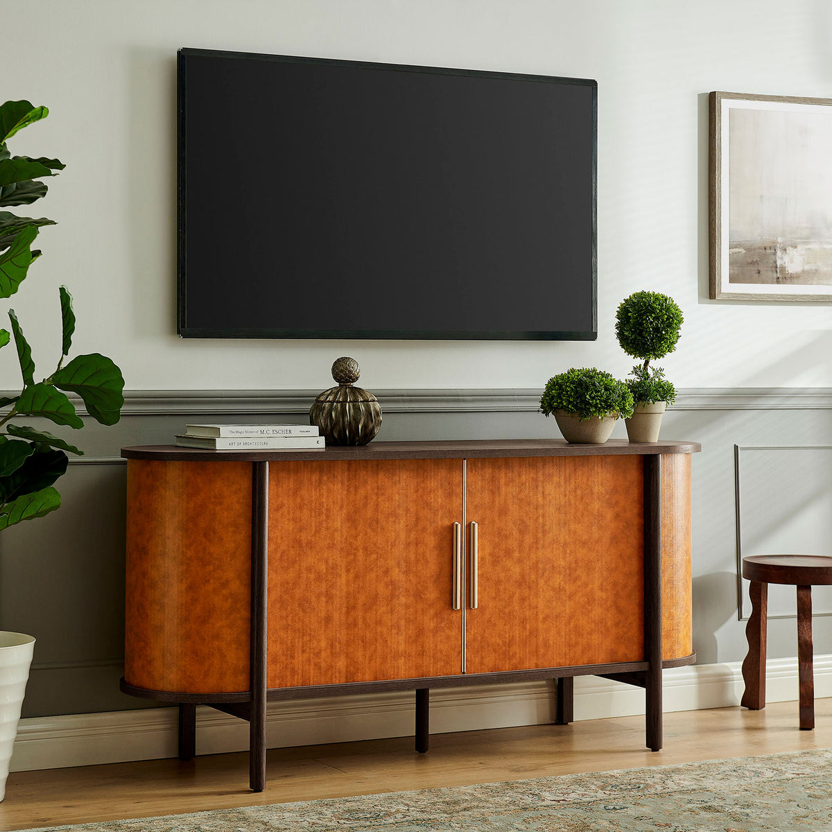 Walnut tan oval sideboard used as a media console beneath a wall-mounted TV with decorative accents || EEI-7373-WAL-TAN || front_angle_room